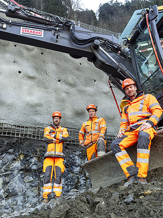 Vier Baufachkräfte vor einem gelben STRABAG Bagger schauen in die Kamera und posieren.