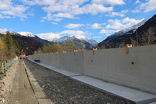 Eine lange, betonierte Stützmauer verläuft entlang einer Strasse. Daneben befindet sich ein vorbereiteter Kiesweg, links liegen Schotter und Material. Im Hintergrund erheben sich schneebedeckte Berge unter einem blauen Himmel mit Wolken.