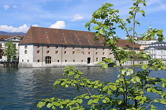 Historisches Landhaus Gebäude mit rotem Ziegeldach direkt an der Aare, mit vielen kleinen Fenstern und roten Fensterläden. Im Vordergrund grüne Zweige eines Baumes, im Hintergrund weitere Altstadtgebäude unter blauem Himmel.