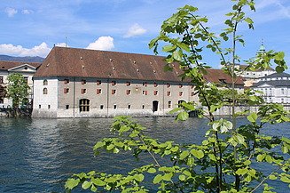 Historisches Landhaus Gebäude mit rotem Ziegeldach direkt an der Aare, mit vielen kleinen Fenstern und roten Fensterläden. Im Vordergrund grüne Zweige eines Baumes, im Hintergrund weitere Altstadtgebäude unter blauem Himmel.