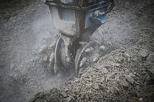 Ein Bagger trägt den Boden ab mit einem Fräserbohrkopf, es staubt 