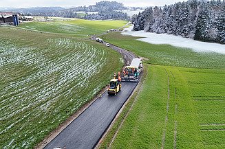 Drohnenaufnahme der K303 mit Asphaltfertiger und einigen Walzen. Grün im Hintergrund und teilweise noch liegendem Schnee.