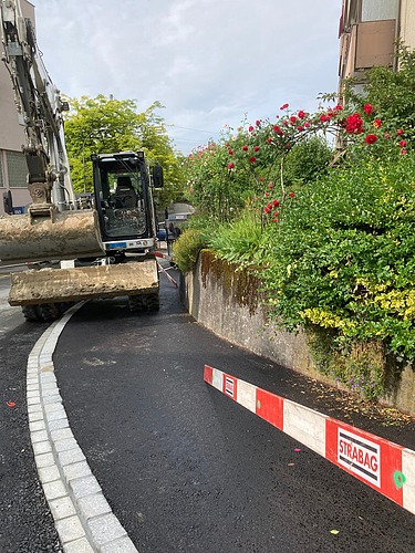 Bagger arbeitet am neu gebauten Trottoir; daneben ist eine rot-weiss markierte Absperrplatte auf der nassen Strasse zu sehen.