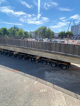 Ein langer Betonbrückenträger liegt auf einem Schwertransportfahrzeug, das auf einer Baustelle steht; im Hintergrund sind Bäume und ein Kran zu sehen.
