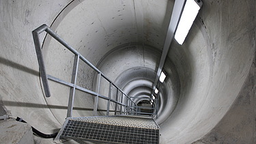 Blick in einen tiefen, zylindrischen Schacht mit Metalltreppe, die Beleuchtung entlang der Wand führt den Blick nach unten.