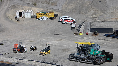Weitläufiges Baustellenareal mit mehreren Baumaschinen, Fahrzeugen und Containern, die Fläche ist von Schotter und Erdmaterial geprägt.