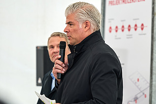 Ein Redner in dunkler Jacke spricht bei einer Veranstaltung in Bürglen in ein Mikrofon. Hinter ihm steht eine weitere Person. Im Hintergrund ist ein Banner der Veranstaltung zu sehen.