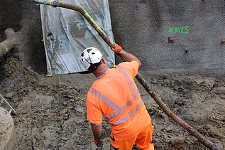 Ein Bauarbeiter mit orangefarbener Arbeitskleidung und weissem Helm hält ein langes Rohr in dem Graben an der Baustelle.