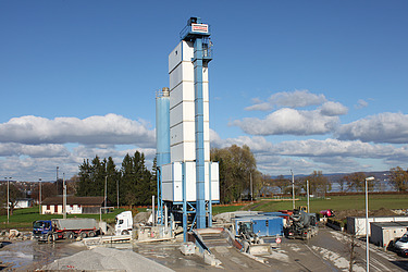 Auf einem Industrieareal steht eine hohe, hellblaue Betonmischanlage, umgeben von Fahrzeugen und Maschinen.