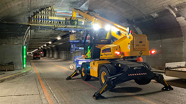  Ein Kran der STRABAG hebt an einem sonnigen Tag ein grosses Brückenelement. Im Hintergrund stehen mehrere Personen auf der Baustelle, neben einem Wohngebäude.