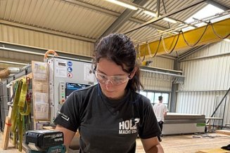 Holzbauhalle im Vordergrund die Zimmerin on Tour mit einem Herz aus Holz in der Hand. Im Hintergrund die CNC-Maschine