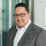 Kaufmännischer Bereichsleiter im Anzug lächelt in die Kamera. Im Hintergrund ist ein Lift zu sehen.