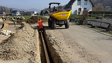  Ein Kran der STRABAG hebt an einem sonnigen Tag ein grosses Brückenelement. Im Hintergrund stehen mehrere Personen auf der Baustelle, neben einem Wohngebäude.
