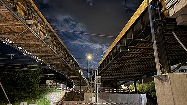  Ein Kran der STRABAG hebt nachts ein langes Brückenteil über Bahngleise. Die Baustelle ist hell erleuchtet.