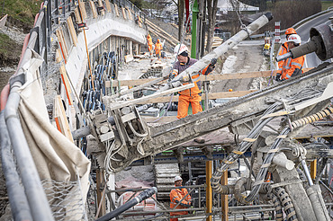 Baurabeiten am Hang - STRABAG Ingenieurbau Nahaufnahme eines Bohrgeräts auf einer Baustelle. Mehrere STRABAG-Arbeiter in orangefarbener Schutzkleidung sind im Hintergrund an einer Betonwand tätig.