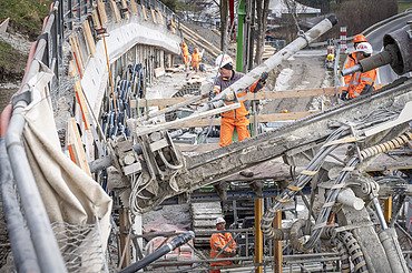 Baurabeiten am Hang - STRABAG Ingenieurbau Nahaufnahme eines Bohrgeräts auf einer Baustelle. Mehrere STRABAG-Arbeiter in orangefarbener Schutzkleidung sind im Hintergrund an einer Betonwand tätig.