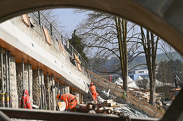 Mehrere STRABAG-Arbeiter in orangefarbener Schutzkleidung sind im Hintergrund an einer Betonwand tätig.