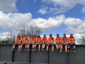 Eine Gruppe von elf Bauarbeitern in orangefarbener Schutzkleidung sitzt nebeneinander auf einem breiten Stahlträger. Hinter ihnen sind Bäume und ein blauer Himmel mit Wolken zu sehen.