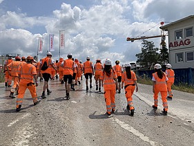 Eine Gruppe von STRABAG-Lernende in orangefarbener Arbeitskleidung und weissen Helmen geht auf einer Baustelle.