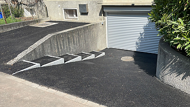  Ein Kran der STRABAG hebt an einem sonnigen Tag ein grosses Brückenelement. Im Hintergrund stehen mehrere Personen auf der Baustelle, neben einem Wohngebäude.