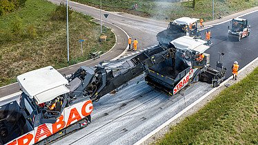 Ein Beschicker, zwei Fertiger, eine Walze und viele Bauarbeiter beim Asphalteinbau 