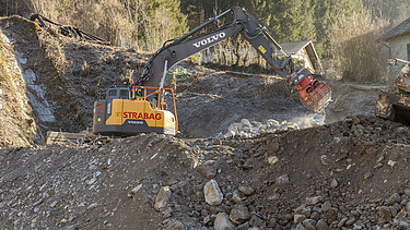 Gelber STRABAG-Bagger von Volvo sortiert mit einem Greifer grosse Steine auf einer Hangbaustelle im Gebirge.