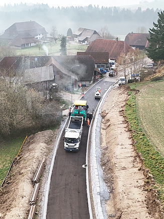Luftaufnahme der Strassenbaustelle im Nebel: Ein Lastwagen liefert Asphalt für den Fertiger auf einer Strasse von Balzenwil. 
