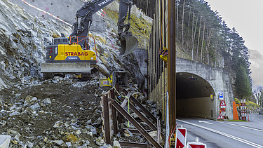 Ein gelber STRABAG Bagger arbeitet an einer Spundwand. Im Hintergrund sieht man einen Tunnel von der Axenstrasse und Bäume von einem Waldgebiet.
