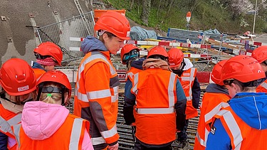 Schulkinder mit Schutzhelmen stehen auf Bewehrungsstahl und beobachten eine Baustelle in Kloten.