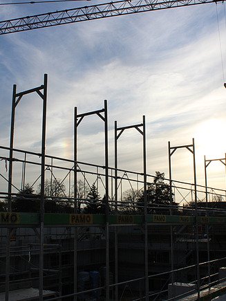 Blick auf eine Baustelle mit mehreren Kränen und Strommasten bei tiefstehender Sonne.