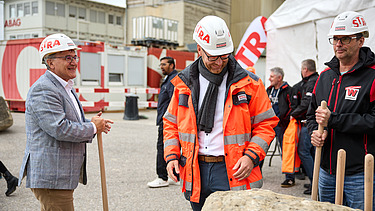 Mehrere Personen stehen bei einem Spatenstich zusammen. Eine Person im grauen Sakko spricht mit einem Arbeiter in orangefarbener Schutzkleidung und Helm, während andere Beteiligte mit Spaten im Hintergrund warten.