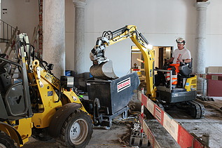 Bauarbeiter in STRABAG-Kleidung bedient einen kleinen Bagger in einem historischen Innenraum mit Säulen. Der Bagger leert Material in einen mit STRABAG-Logo versehenen Container. Links im Bild ist eine gelbe Baumaschine zu sehen. Die Baustelle ist mit Absperrbarrieren gesichert.