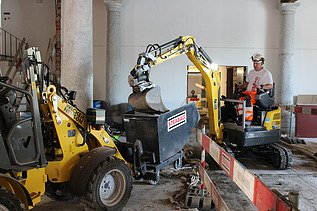 Bauarbeiter in STRABAG-Kleidung bedient einen kleinen Bagger in einem historischen Innenraum mit Säulen. Der Bagger leert Material in einen mit STRABAG-Logo versehenen Container. Links im Bild ist eine gelbe Baumaschine zu sehen. Die Baustelle ist mit Absperrbarrieren gesichert.