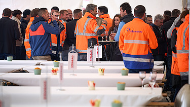 Viele Besucherinnen und Besucher stehen in einem grossen Festzelt beisammen. Mehrere Personen tragen orange Sicherheitswesten von STRABAG. Im Vordergrund sind gedeckte Tische mit Dekoration zu sehen.
