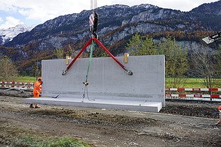 Eine schwere Betonelementwand hängt an roten und grünen Hebegurten unter dem Kranhaken. Auf der Baustelle stehen Mitarbeitende in orangefarbener Schutzausrüstung daneben, während im Hintergrund eine Berglandschaft mit bewaldeten Hängen zu sehen ist.
