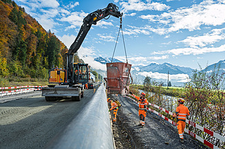 Ein Bagger hebt einen grossen Betonbehälter über eine Strassenbaustelle mehrere Arbeiter in oranger Schutzkleidung stehen daneben und sichern die Arbeiten. Im Hintergrund sind Herbstbäume zu sehen.