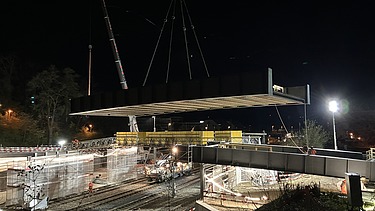 Bei Nacht wird ein grosses Brückenelement mit zwei Pneukranen über die Bahnlinie gehoben. Unterhalb der schwebenden Brücke sind die beleuchteten Gleise und Baustellenbereiche zu sehen. Mehrere Arbeiter sind auf der Baustelle im Einsatz.