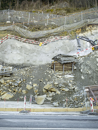 Baugrube Los 105 der neuen N4 Axenstrasse in Sisikon im Ganzen. Gelber STRABAG Bagger steht in der Baugrube und spitzt Felsgestein ab. 