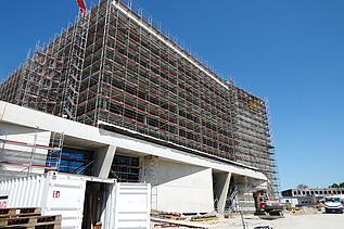 Nahaufnahme eines grossen, eingerüsteten Gebäudes auf einer Baustelle. Die Fassade ist vollständig von Gerüsten umgeben, im Vordergrund stehen ein geöffneter STRABAG Baucontainer, Baupaletten und eine Baumaschine. Rechts im Bild ein weisser Lieferwagen und ein im Bau befindliches Nebengebäude, darüber ein klarer blauer Himmel.