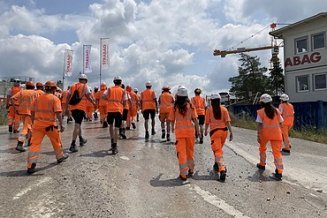 Eine Gruppe von STRABAG-Lernende in orangefarbener Arbeitskleidung und weissenHelmen geht auf einer Baustelle.