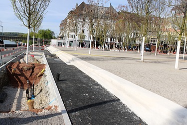 Eine Baustelle im Stadtgebiet: Arbeiter gestalten Wege und Grünflächen neu. Im Vordergrund ist frischer Asphalt zu sehen, daneben gepflanzte Bäume.