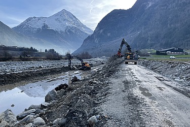 Zwei STRABAG Bagger arbeiten in einem gesicherten Bach. Im Hintergrund sieht macht die Schweizer Berge.
