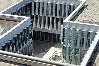 Blick von oben auf den Innenhof des modernen Neubaus mit markanter Fassade: Die Glasflächen sind durch dreieckig zulaufende, vertikal angeordnete Fassadenelemente rhythmisch strukturiert. Die Geometrie erzeugt ein prägnantes Licht- und Schattenspiel. Im Innenhof ist ein heller Betonboden mit einem abgesperrten Treppenabgang zu sehen.