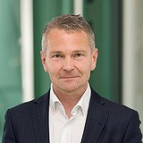 Technischer Direktionsleiter im Anzug lächelt in die Kamera. Im Hintergrund ist ein Lift zu sehen.