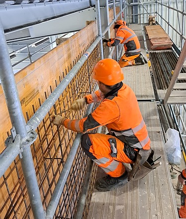  Zwei Bauarbeiter in orangefarbener Warnkleidung von STRABAG arbeiten auf einem Gerüst an einer Bewehrung.