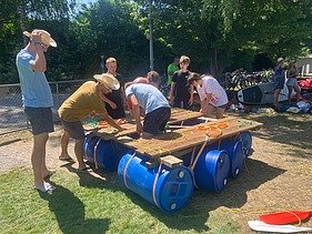 Lernende in Alltagskleider bauen ein Floss bei strahlendem Sonnenschein.