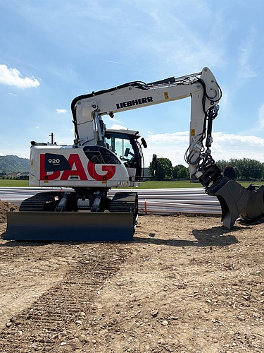Ein weisser STRABAG Liebherr-Raupenbagger steht einsatzbereit auf einer Erd- und Kiesfläche vor einem Baucontainer.