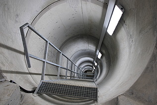 Blick in einen tiefen, zylindrischen Schacht mit Metalltreppe, die Beleuchtung entlang der Wand führt den Blick nach unten.