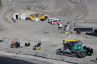 Weitläufiges Baustellenareal mit mehreren Baumaschinen, Fahrzeugen und Containern, die Fläche ist von Schotter und Erdmaterial geprägt.