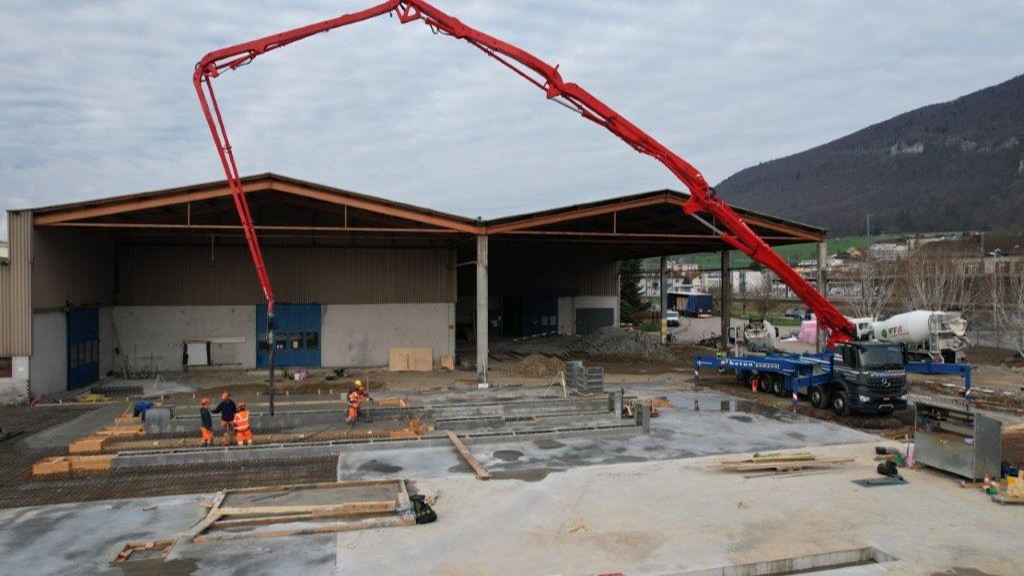 Die Baustelle «Erweiterung Bourquin» mit einem grossen, roten Betonpumpen-Lastwagen, der Beton für die Gebäudeerweiterung liefert.